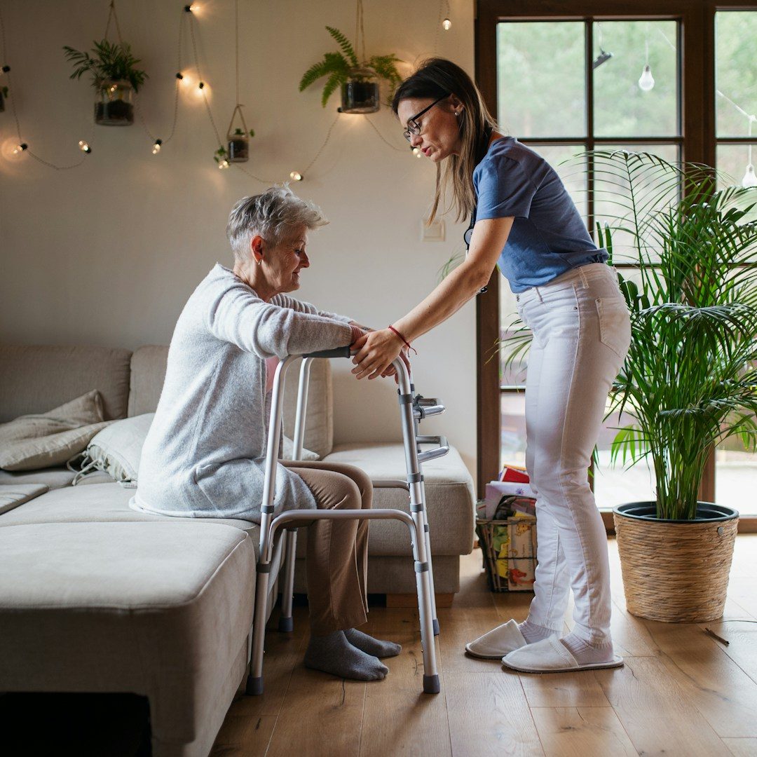 Eine Betreuungskraft unterstützt eine Seniorin im Wohnzimmer liebevoll beim Aufstehen mit einer Gehhilfe.