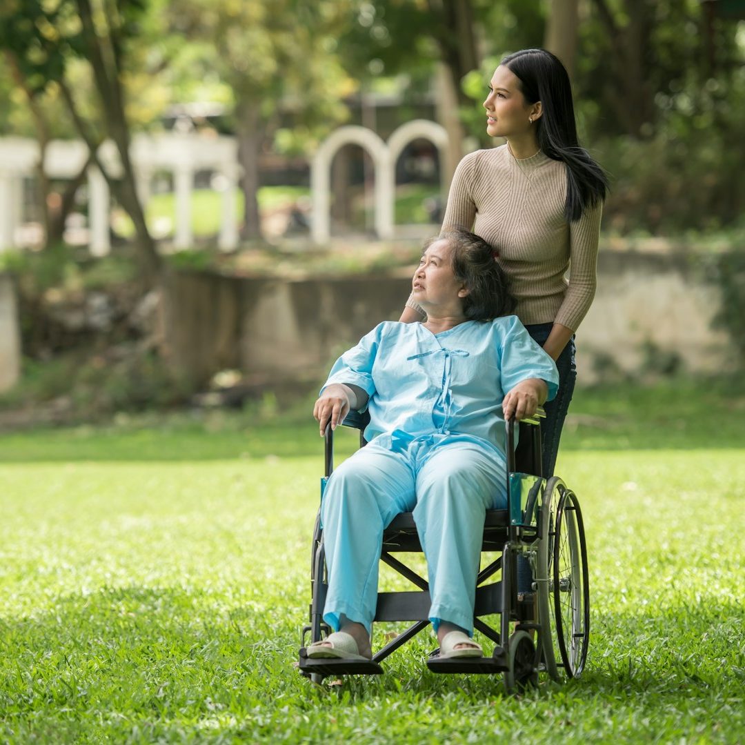 Eine aufmerksame Betreuungskraft begleitet eine Seniorin im Rollstuhl bei einem entspannten Spaziergang im Park.
