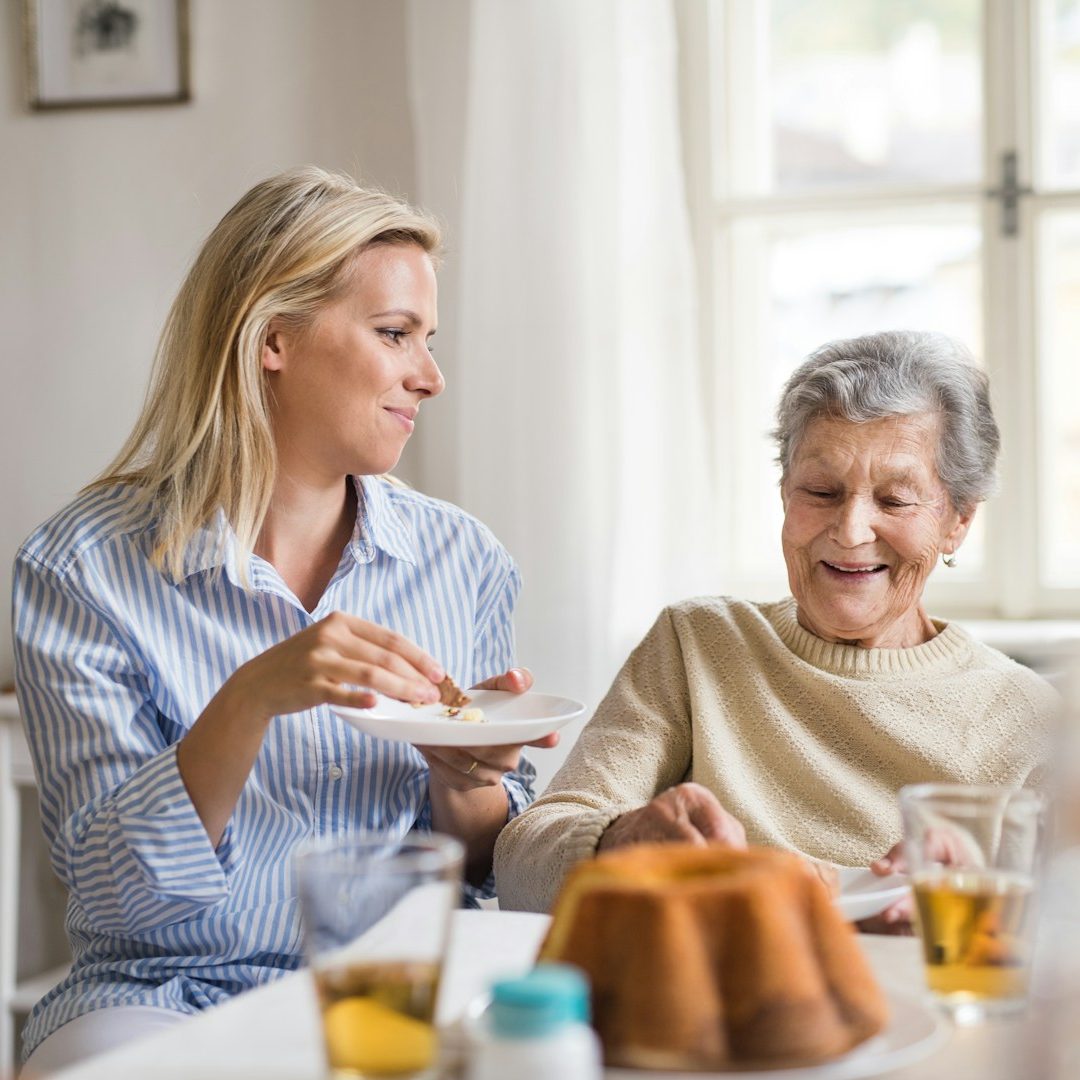 Eine freundliche junge Betreuungskraft teilt einen geselligen Moment beim Kaffeetrinken mit einer älteren Dame, die fröhlich lächelt.
